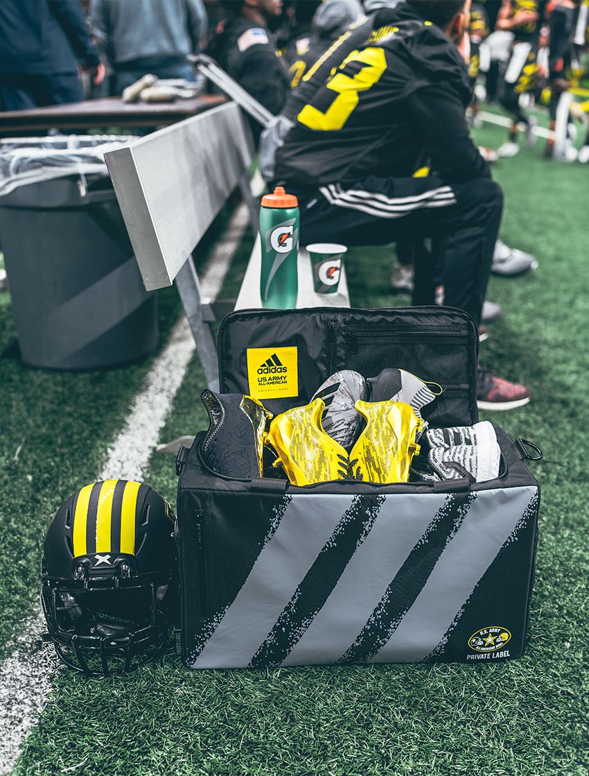 Black bag with 3 yellow stipes in a football arena with a football player sitting behind the bag , cleats packed inside