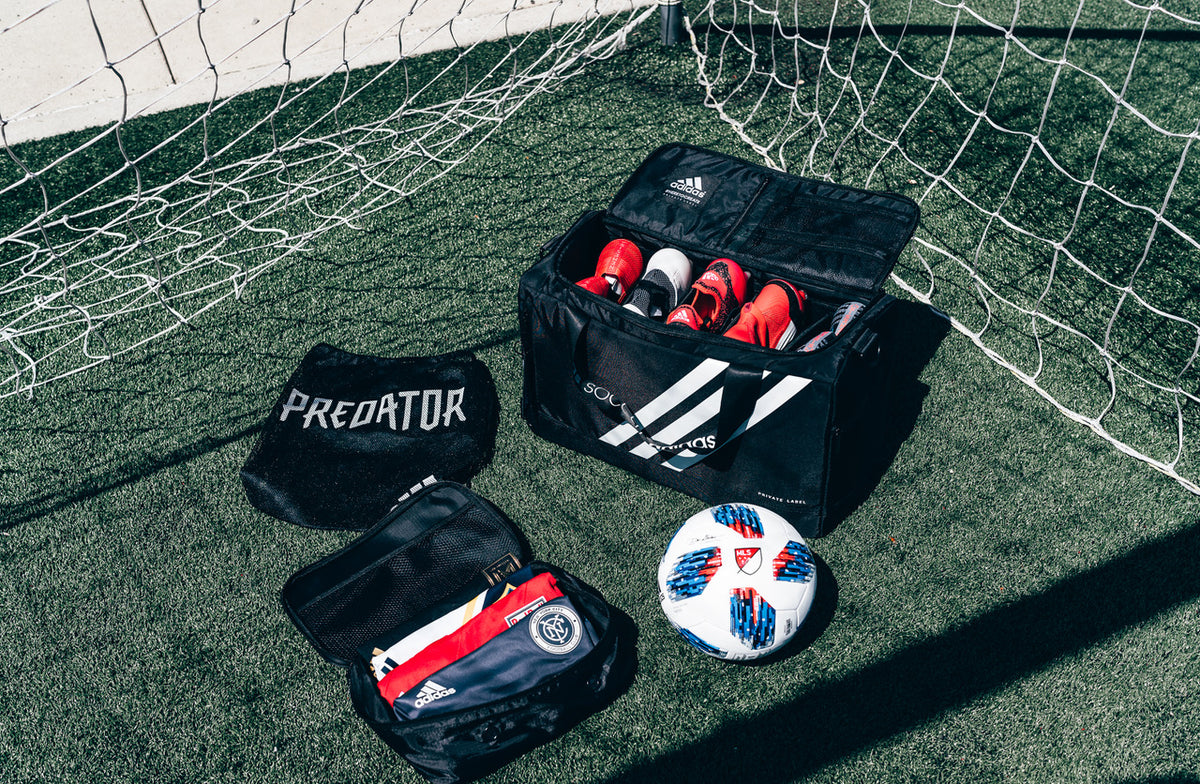 Adidas soccer gear including shoes, balls, and bags on a grass field with a goal net in the background.