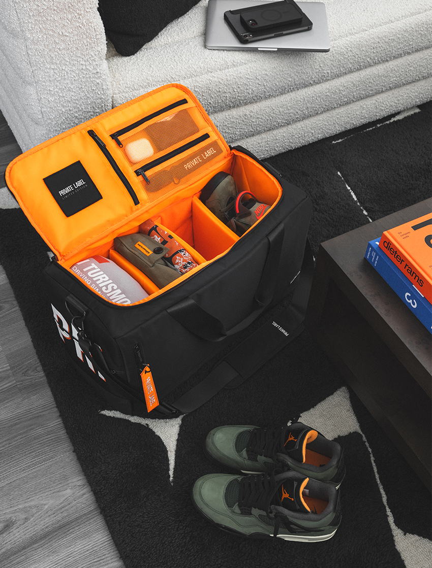 A black and bright orange travel organizer bag is shown open on a dark rug in a living room setting, filled with shoes and accessories. A pair of green and black sneakers sits nearby, with a sofa and side table visible in the background.