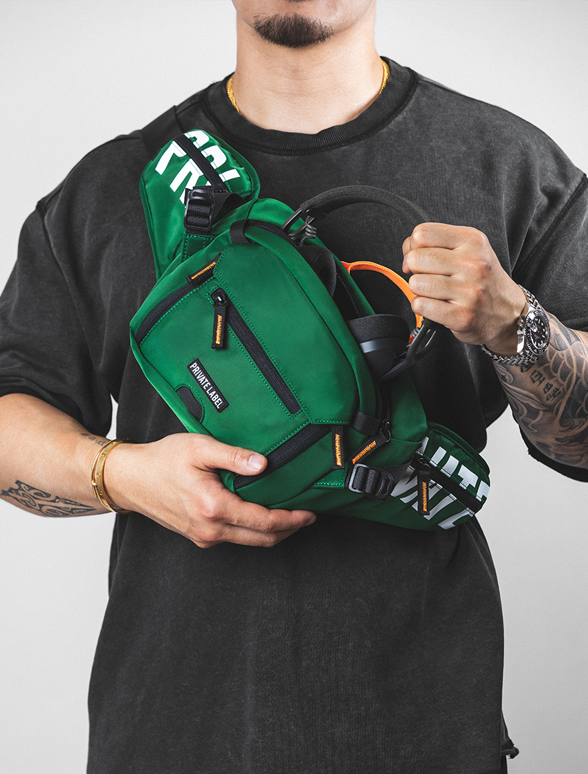 A man wearing a black t-shirt holds a green  sling bag across his chest. The bag features black straps, white text on the side, and orange zipper pulls, shown against a plain light gray background.