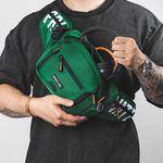 A man wearing a black t-shirt holds a green  sling bag across his chest. The bag features black straps, white text on the side, and orange zipper pulls, shown against a plain light gray background.