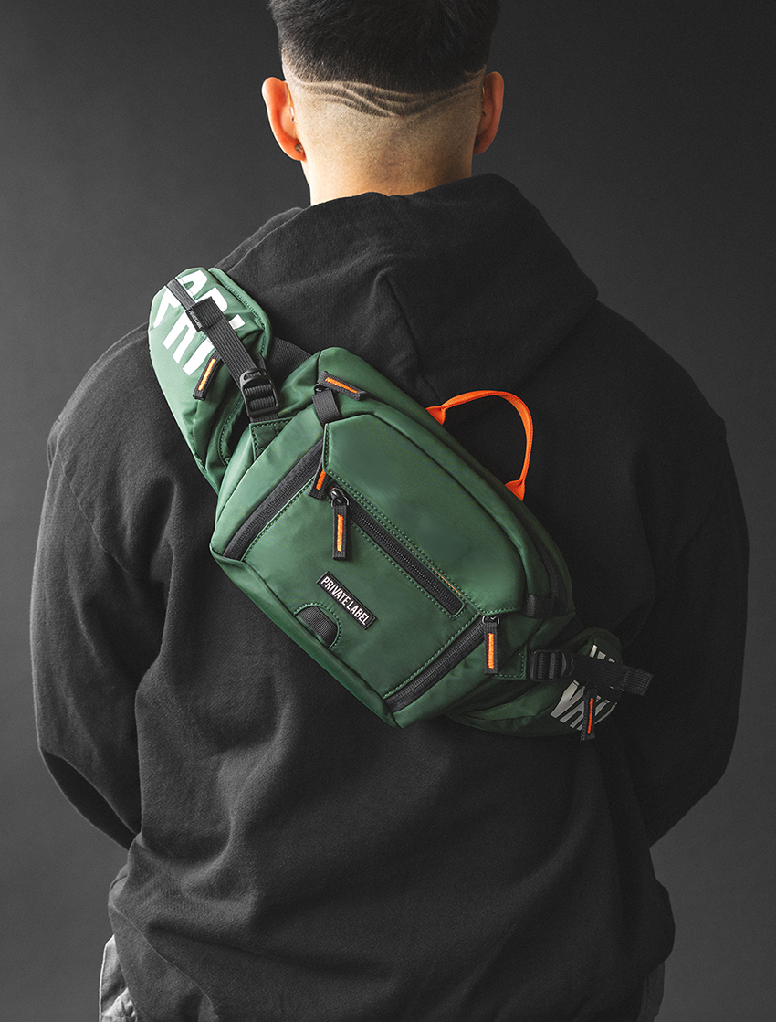 A man viewed from behind wearing a dark green crossbody sling bag with orange accents and black straps over a black hoodie against a dark gray studio background.