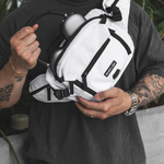 A white crossbody sling bag with black straps and zippers, worn across the chest of a person wearing a dark gray t-shirt. The bag has a small black label on the front pocket that reads 'PRIVATE LABEL'.