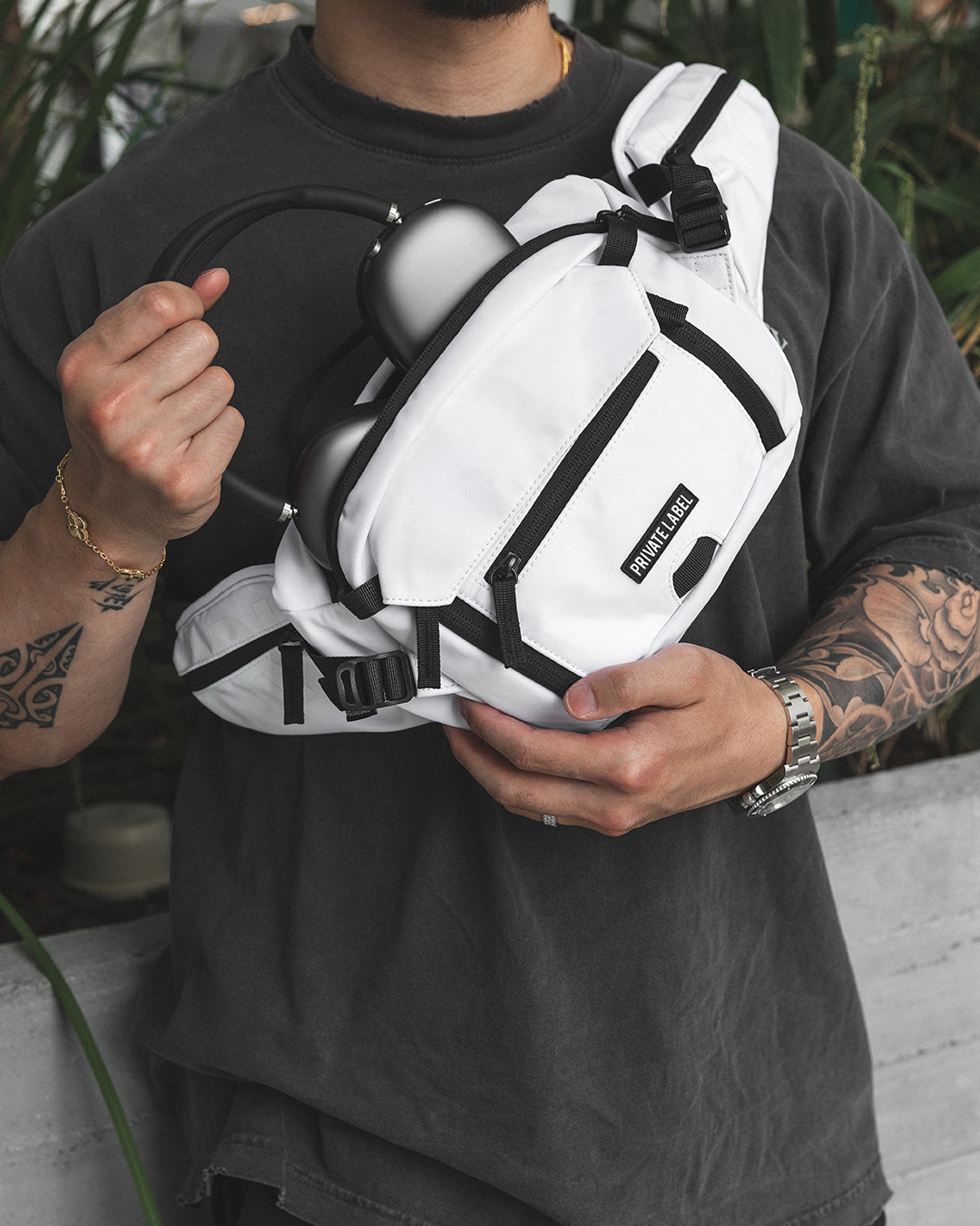 A white crossbody sling bag with black straps and zippers, worn across the chest of a person wearing a dark gray t-shirt. The bag has a small black label on the front pocket that reads 'PRIVATE LABEL'.