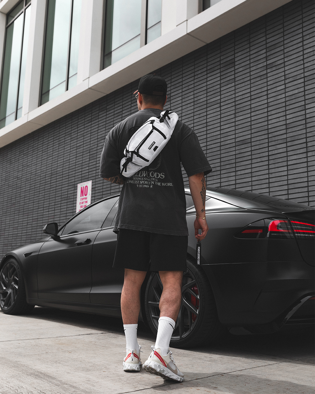 A man viewed from behind wearing a white and black crossbody sling bag over a black t-shirt, standing next to a black car in front of a modern gray brick building.