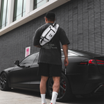 A man viewed from behind wearing a white and black crossbody sling bag over a black t-shirt, standing next to a black car in front of a modern gray brick building.