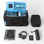 A black carry-on luggage duffle set shown open on a light gray studio background, with a bright blue interior organizer panel, packed accessories inside, a separate black zippered pouch, headphones, a cylindrical bottle, a passport, and folded clothing arranged around it.
