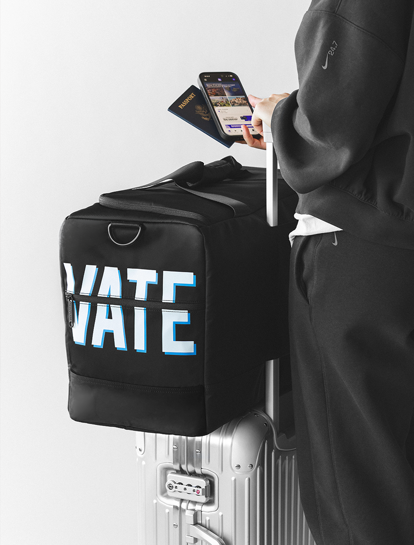 A black duffel bag with large white and blue 'VATE' lettering is attached to a silver rolling suitcase. A person in a dark hoodie holds a phone and passport beside the bag, against a clean light gray studio background.