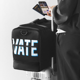 A black duffel bag with large white and blue 'VATE' lettering is attached to a silver rolling suitcase. A person in a dark hoodie holds a phone and passport beside the bag, against a clean light gray studio background.
