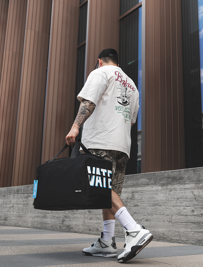 A man from behind wearing a white graphic t-shirt, camouflage shorts, and white sneakers, carrying a large black duffel bag with 'VATE' printed on the side, walking outdoors in front of a modern building with brown vertical siding.