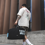 A man from behind wearing a white graphic t-shirt, camouflage shorts, and white sneakers, carrying a large black duffel bag with 'VATE' printed on the side, walking outdoors in front of a modern building with brown vertical siding.