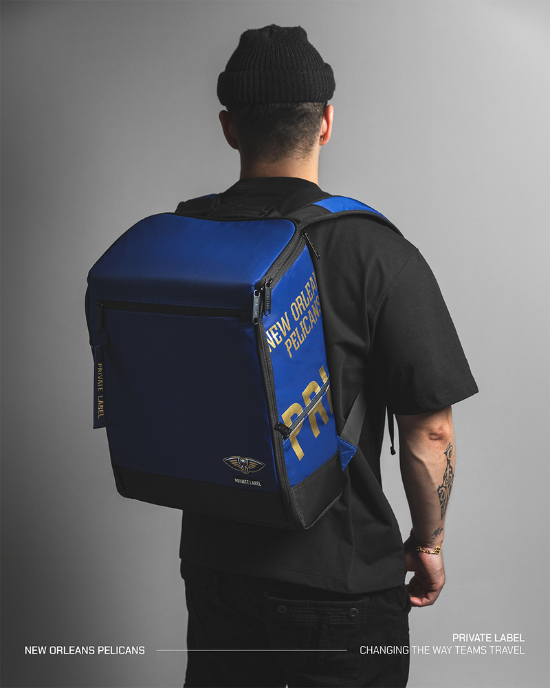 A man viewed from behind wearing a black beanie, black t-shirt, and black pants, carrying a royal blue and black New Orleans Pelicans private label backpack. The backpack features gold and white lettering and a small team logo.