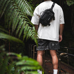 Person wearing a white t-shirt, black shorts, and white sneakers with a black sling bag in a forest setting.