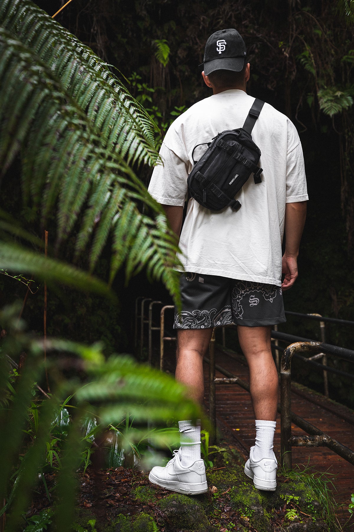 Person wearing a white t-shirt, black shorts, and white sneakers with a black sling bag in a forest setting.