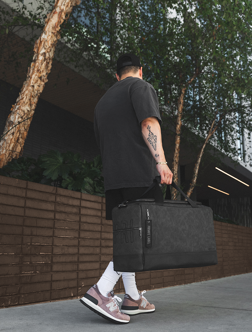 A man carrying a large black duffel bag with a textured pattern, walking outdoors next to a modern building with trees and a brick wall.
