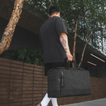 A man carrying a large black duffel bag with a textured pattern, walking outdoors next to a modern building with trees and a brick wall.