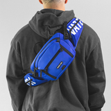 A person wearing a gray hoodie has their back turned to the camera, carrying a blue waist/sling bag with white branding and black straps.