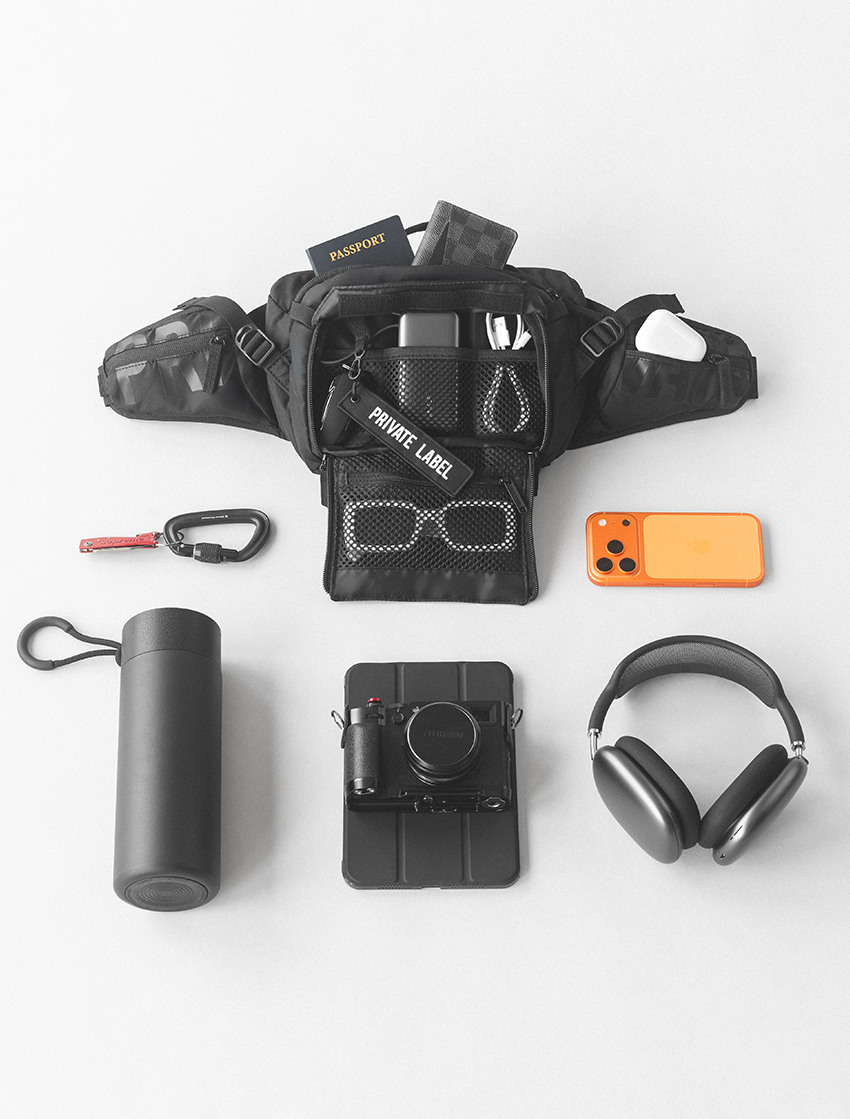 black waist bag with tech accessories, wallet and sunglasses surrounded by a bottle , tablet , headphones &  phone on a light gray background