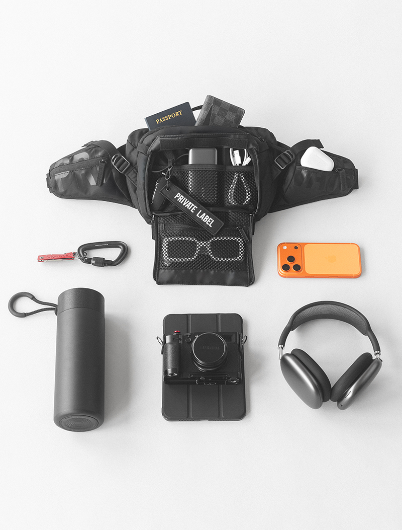 black waist bag with tech accessories, wallet and sunglasses surrounded by a bottle , tablet , headphones &  phone on a light gray background