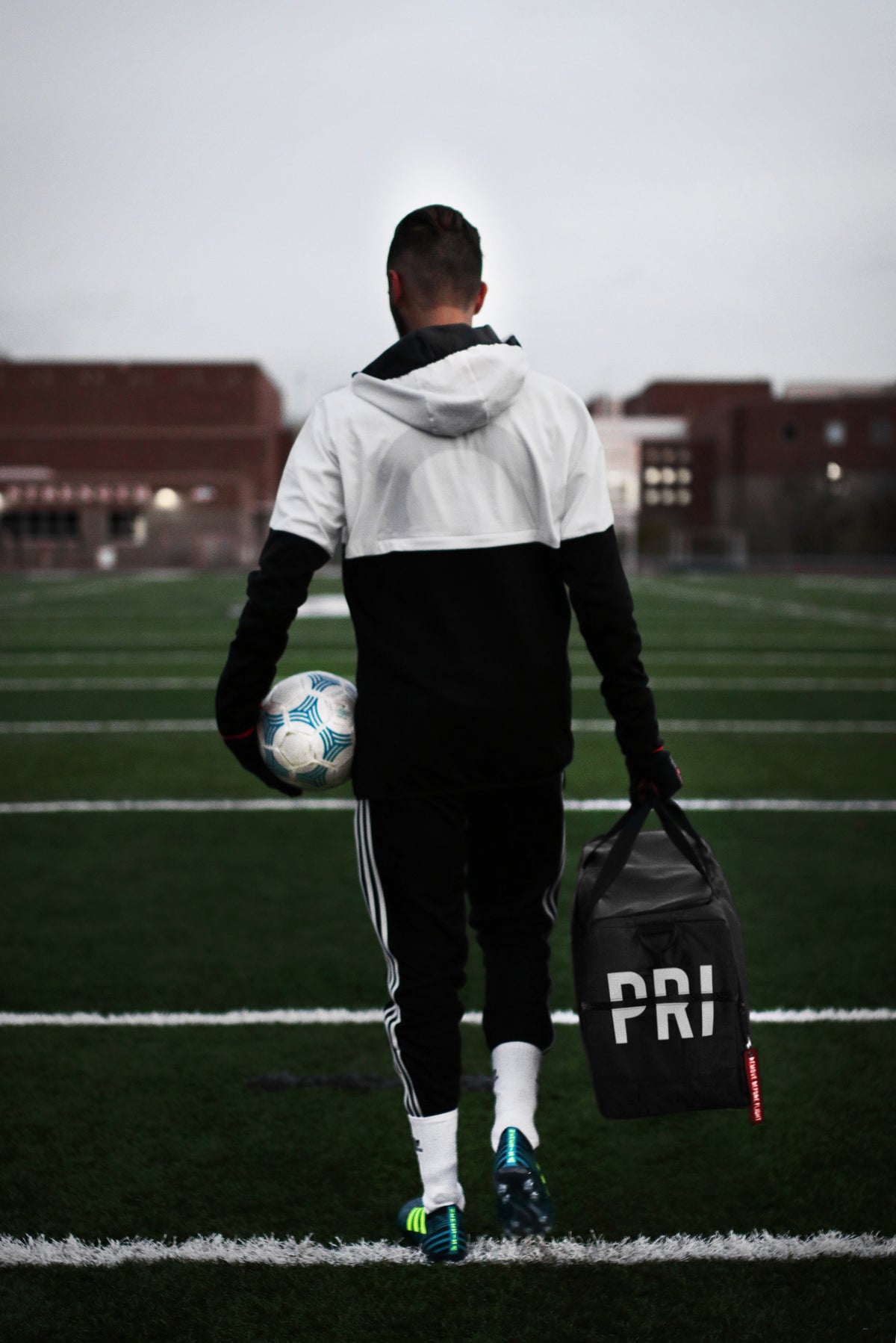 Person walking on a sports field with a soccer ball and a bag labeled 'PRI'.