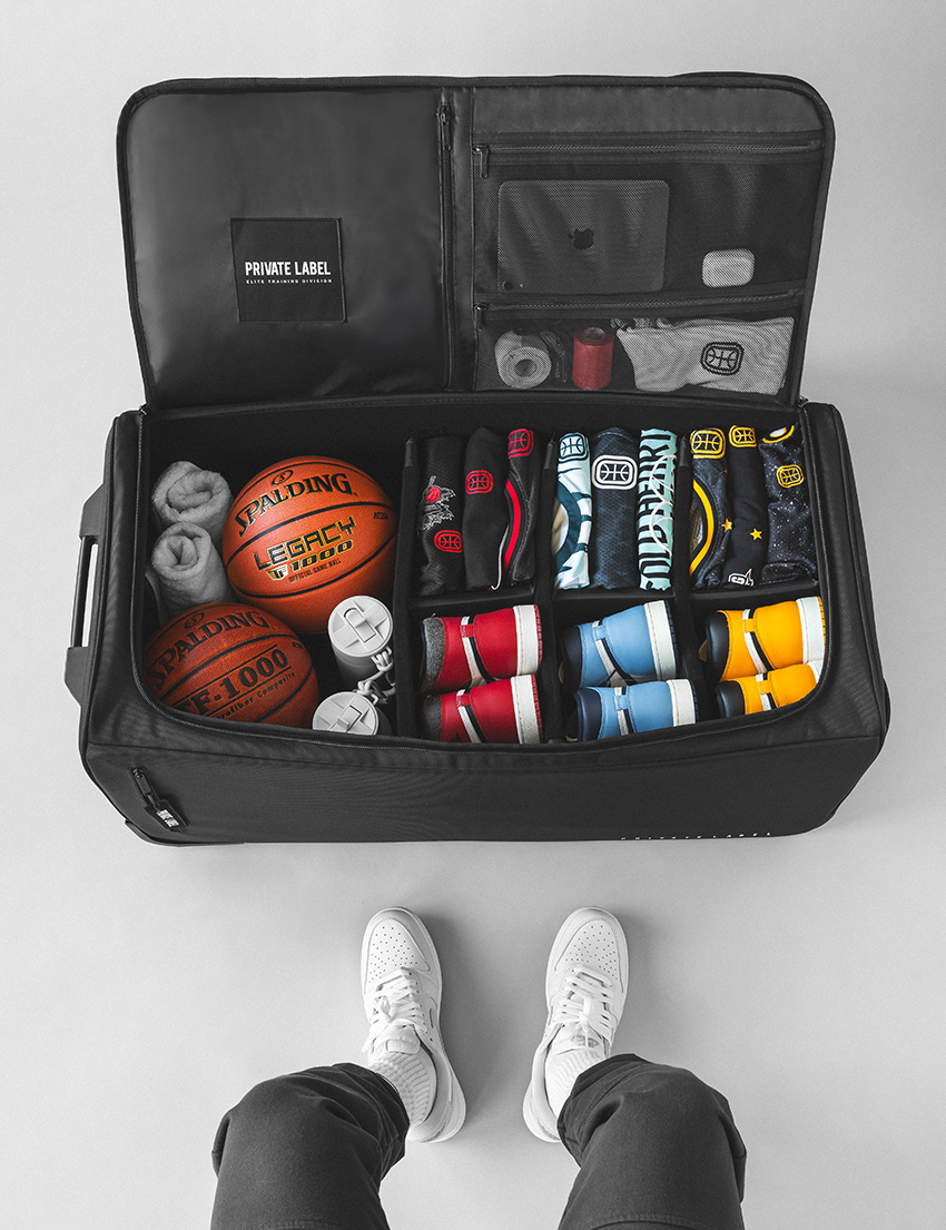 interior view of a black suit case with basketball gear loaded inside on a light gray background and a model sitting near by
