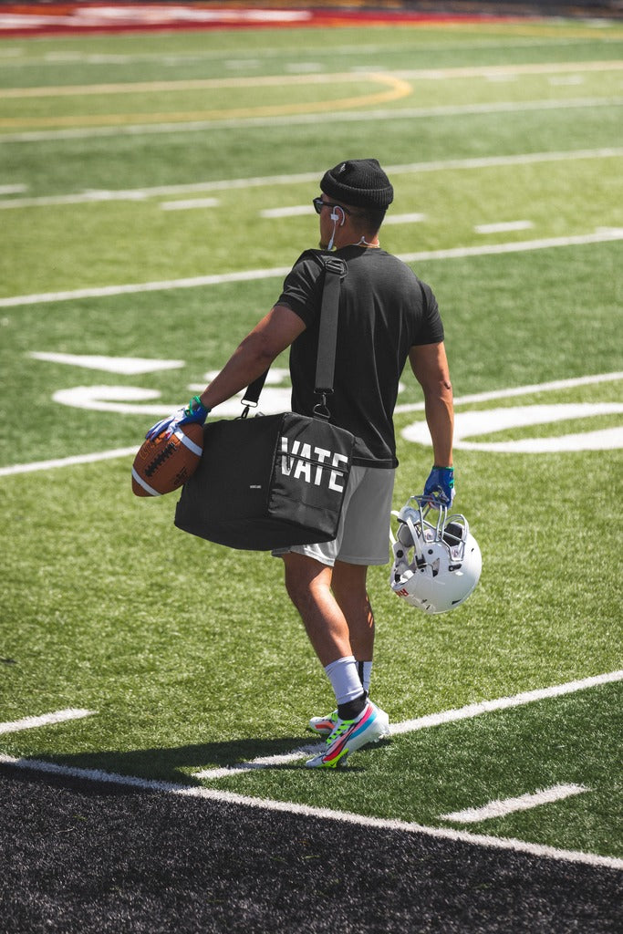 football player walking on the football field with a bag showing "VATE" and helmet 